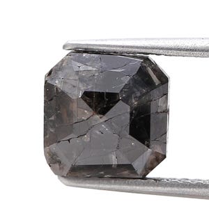 May include: A faceted, rectangular-cut black diamond held by a pair of silver tweezers. The gemstone has a complex, multi-faceted surface with visible internal fractures, showcasing its natural characteristics. The diamond's dark color and geometric shape are prominent.