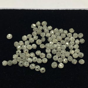 May include: A collection of small, faceted, light gray diamonds. Each diamond has a slightly irregular shape and a small hole drilled through it. The diamonds are scattered on a black surface.
