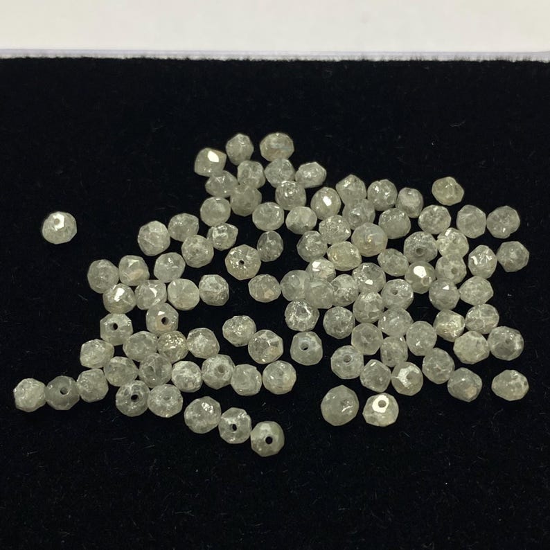 May include: A collection of small, faceted, light gray diamonds. Each diamond has a slightly irregular shape and a small hole drilled through it. The diamonds are scattered on a black surface.