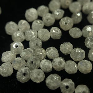 May include: A collection of faceted, clear crystal beads with small holes. The beads are irregularly shaped and reflect light, creating a sparkling effect. The beads are scattered on a dark surface, suggesting jewelry making supplies.