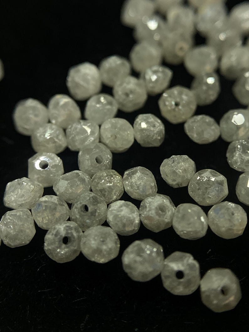 May include: A collection of faceted, clear crystal beads with small holes. The beads are irregularly shaped and reflect light, creating a sparkling effect. The beads are scattered on a dark surface, suggesting jewelry making supplies.