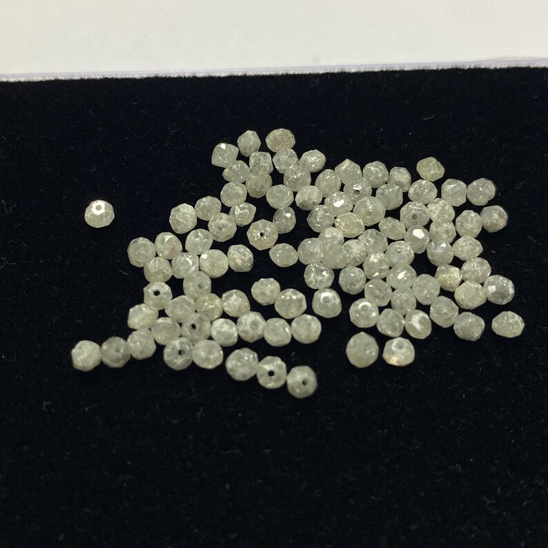May include: A collection of small, faceted, light-colored beads, possibly gemstones or crystals, scattered on a dark surface. The beads have a slightly irregular shape and appear to be translucent. The image is a close-up, highlighting the texture and detail of the beads.