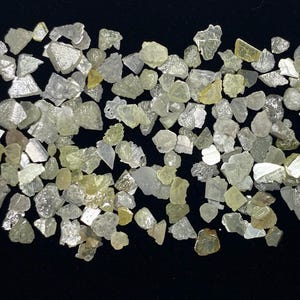 Op de afbeelding: Een verzameling ruwe diamanten in verschillende vormen en maten. De edelstenen vertonen een reeks kleuren, waaronder helder, lichtgeel en grijs, tegen een zwarte achtergrond. Deze ruwe diamanten zijn waarschijnlijk voor het maken van sieraden.