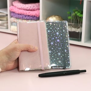 May include: Two decorative notebooks with a pink and a grey cover. The grey cover has a holographic star pattern. The notebooks are closed and lying on a pink surface.