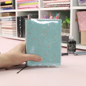 May include: A turquoise blue planner with a clear plastic cover. The planner has a repeating pattern of silver glitter stars. The planner has the text "2024" and "hi-line" printed on the front cover.