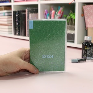 May include: A green glitter-covered planner with a clear plastic cover. The planner has the year "2024" printed on the front.