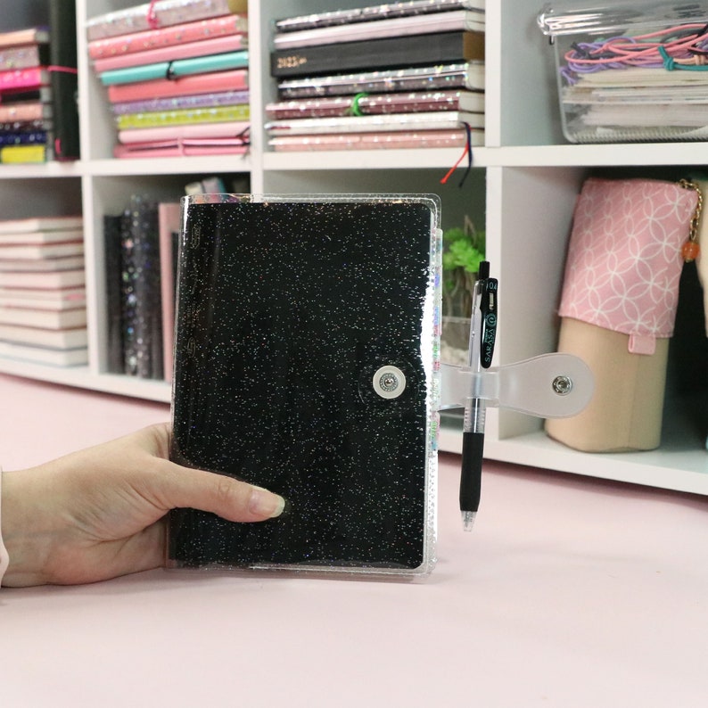 May include: A clear plastic binder with a black glitter cover. The binder has a silver button closure and a pen holder on the side. A black pen is inserted in the pen holder.