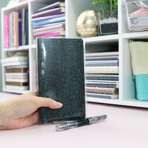 May include: A rectangular notebook with a dark, glittery black cover encased in a clear plastic sleeve, positioned next to a transparent fountain pen with iridescent metallic accents on a smooth, light pink surface.