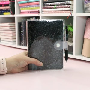 May include: A clear plastic cover with a black glitter notebook inside. The notebook has a silver button closure and a black pen is tucked into the cover.