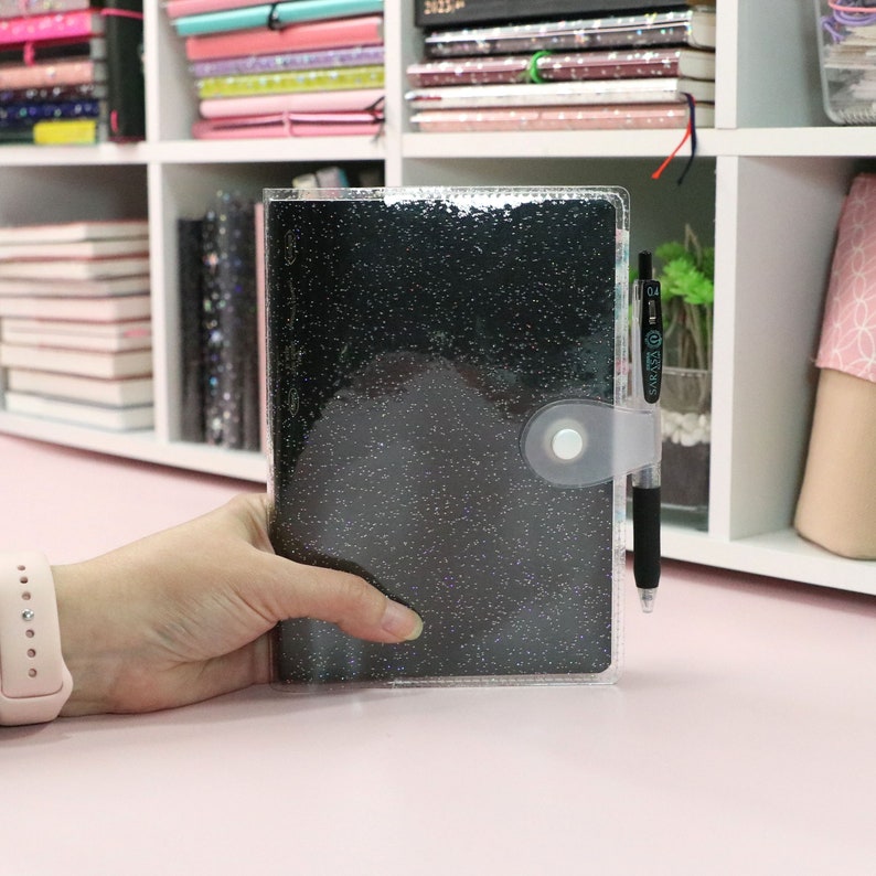 May include: A clear plastic cover with a black glitter notebook inside. The notebook has a silver button closure and a black pen is tucked into the cover.