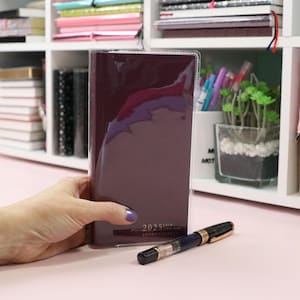May include: A burgundy leather-bound planner with a clear plastic cover. The planner has the year "2025" printed on the front in gold lettering.