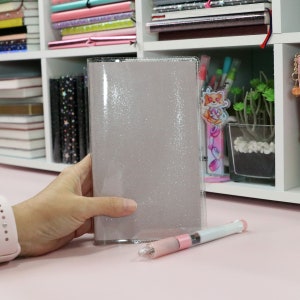 May include: A clear, glitter-covered notebook cover with a silver shimmer. The cover is held in a hand and a pink pen is lying on a pink surface.