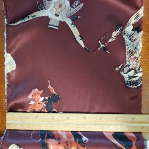 May include: Burgundy fabric with a repeating pattern of cowboys riding horses and a stylized Native American design. The fabric is smooth and has a satin finish.