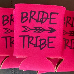May include: Six pink can coolers with black text and an arrow design. The text on the coolers reads "Bride Tribe".