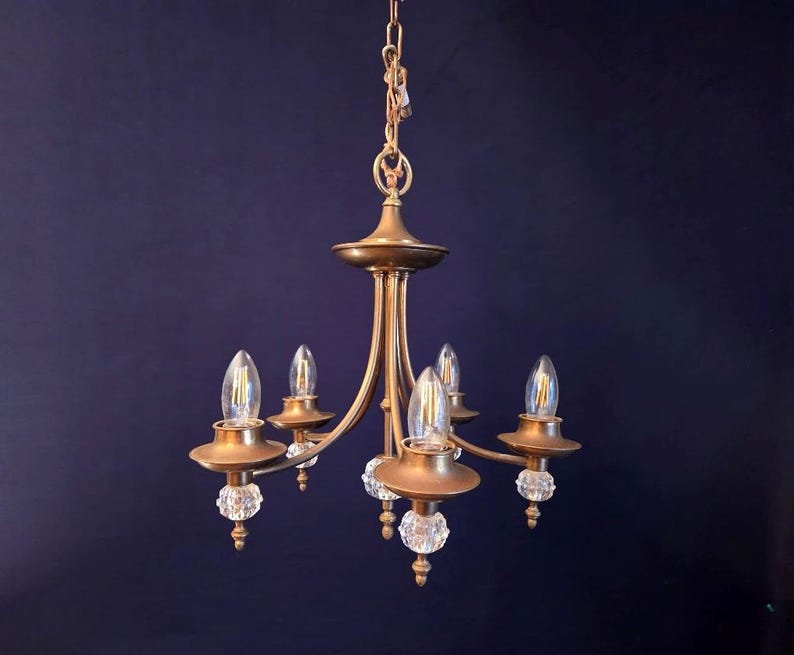 May include: A vintage brass chandelier with five lights. The fixture has a central column with curved arms extending outwards, each holding a light bulb. Clear crystal ball accents are visible. The chandelier hangs from a chain.