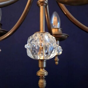May include: Close-up of a vintage chandelier with a brass frame and clear, faceted glass accents. The chandelier has multiple arms with light bulbs and decorative glass elements. The background is a dark blue.