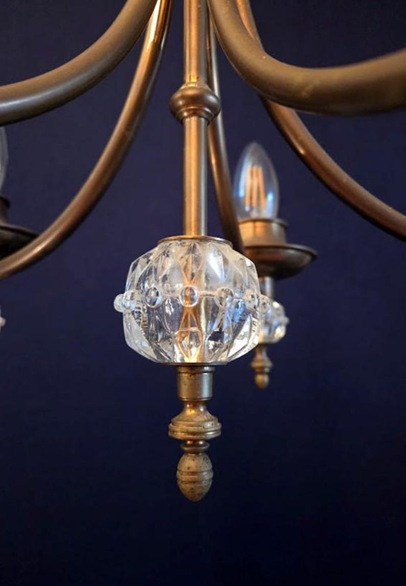 May include: Close-up of a vintage chandelier with a brass frame and clear, faceted glass accents. The chandelier has multiple arms with light bulbs and decorative glass elements. The background is a dark blue.