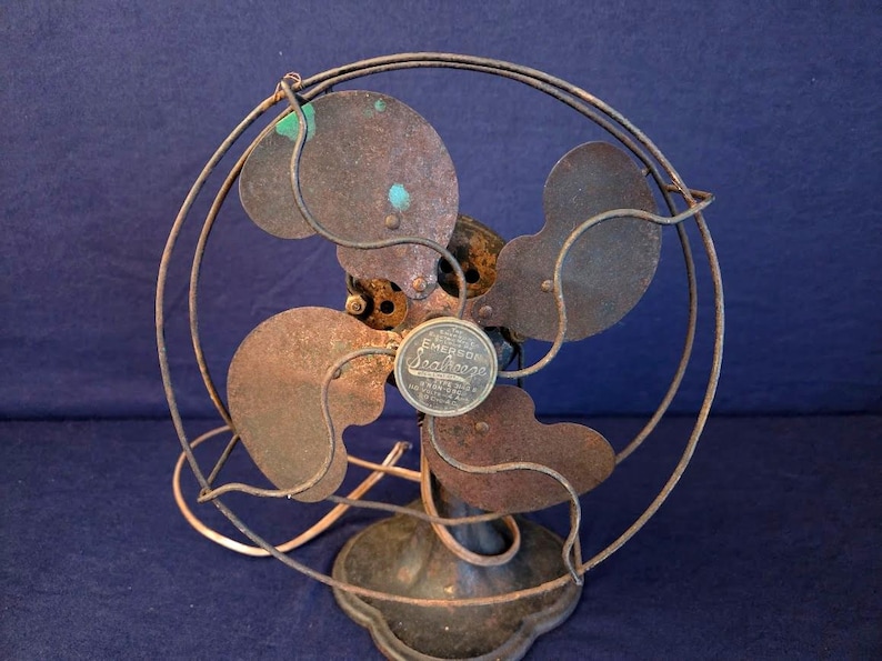 Peut inclure: Un ancien ventilateur Emerson Seabreeze avec une cage circulaire et quatre pales. Le ventilateur pr&eacute;sente un aspect rouill&eacute; et vieilli, avec une base en m&eacute;tal et un petit embl&egrave;me circulaire au centre. Objet de d&eacute;coration vintage.