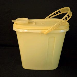 May include: Pale yellow plastic container with a lid and handle. The container has a rectangular shape with a slightly tapered design. The lid has a small spout and the handle has a series of oval cutouts.