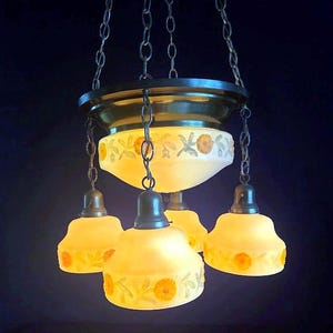 May include: Antique chandelier with four yellow glass shades, each adorned with orange floral patterns. The fixture is suspended by chains from a bronze-colored canopy. The shades are a soft yellow color, and the overall design is vintage.