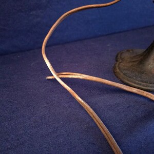 Peut inclure: Gros plan sur une base de ventilateur antique et un cordon d'alimentation. La base est en m&eacute;tal fonc&eacute; avec un bord d&eacute;coratif. Le cordon est marron et semble vieilli. Le fond est un tissu bleu fonc&eacute;.