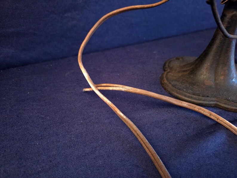 Peut inclure: Gros plan sur une base de ventilateur antique et un cordon d'alimentation. La base est en m&eacute;tal fonc&eacute; avec un bord d&eacute;coratif. Le cordon est marron et semble vieilli. Le fond est un tissu bleu fonc&eacute;.