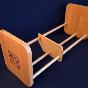 May include: A light-colored wooden book stand with a rectangular base and two square supports connected by three dowels. The base features a stamped logo. The stand is designed to hold books upright.