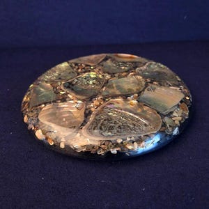 May include: A round, decorative object with a dark, glossy border. The center is filled with iridescent, shell-like pieces and small, light-colored pebbles. The overall effect is a natural, textured design.