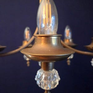May include: Close-up of a vintage brass chandelier with clear glass light bulbs. The chandelier has a decorative crystal glass element at the base of each light fixture. The background is a dark blue.