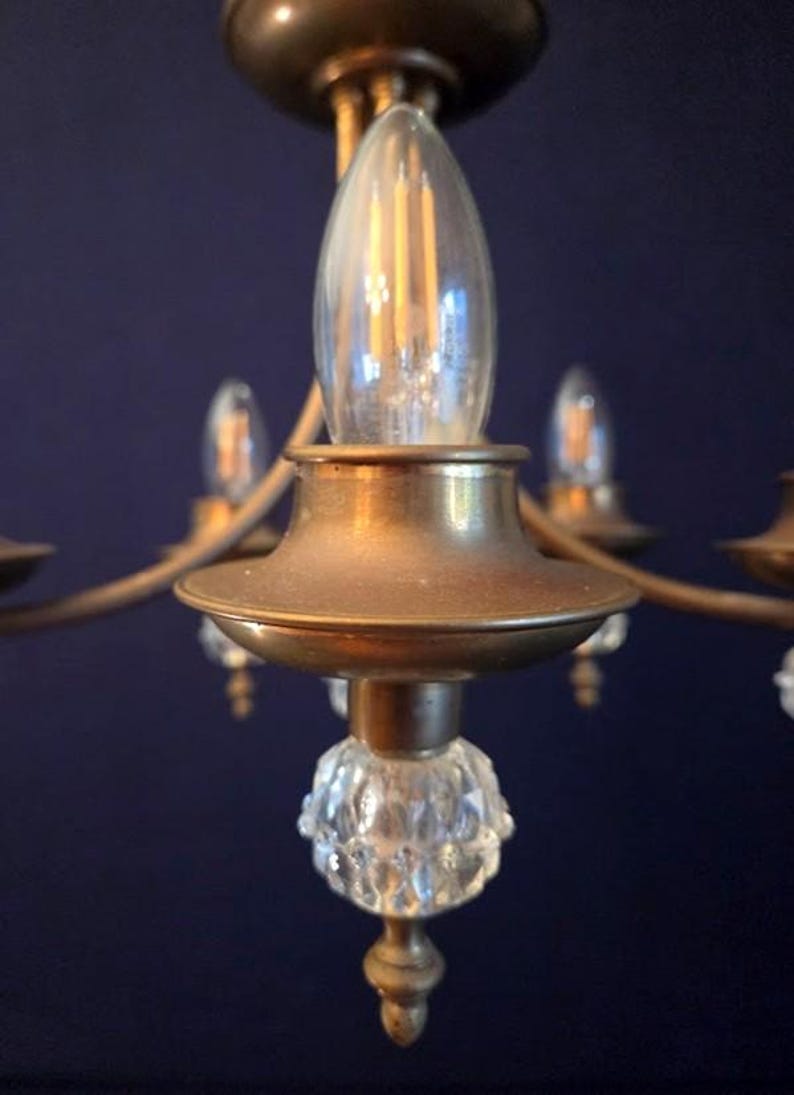 May include: Close-up of a vintage brass chandelier with clear glass light bulbs. The chandelier has a decorative crystal glass element at the base of each light fixture. The background is a dark blue.