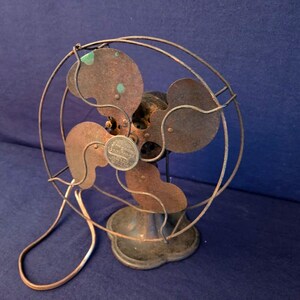 Peut inclure: Un ventilateur &eacute;lectrique ancien avec une cage circulaire et quatre pales. Le ventilateur a une apparence rouill&eacute;e et vieillie avec une finition de couleur cuivre. La base est ronde et le ventilateur est pos&eacute; sur un fond bleu fonc&eacute;.