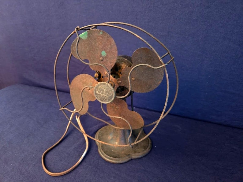 Peut inclure: Un ventilateur &eacute;lectrique ancien avec une cage circulaire et quatre pales. Le ventilateur a une apparence rouill&eacute;e et vieillie avec une finition de couleur cuivre. La base est ronde et le ventilateur est pos&eacute; sur un fond bleu fonc&eacute;.