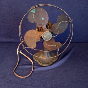 Peut inclure: Un ancien ventilateur &eacute;lectrique rouill&eacute;. Il poss&egrave;de quatre pales prot&eacute;g&eacute;es par une cage m&eacute;tallique et un socle rond. Le cordon d'alimentation est visible. Les composants m&eacute;talliques montrent des signes d'usure, t&eacute;moignant de son anciennet&eacute;.