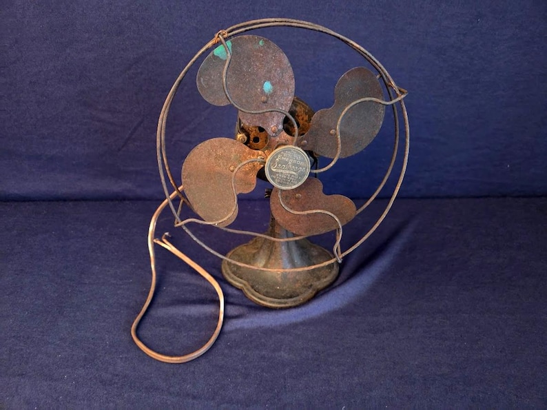 Peut inclure: Un ancien ventilateur &eacute;lectrique rouill&eacute;. Il poss&egrave;de quatre pales prot&eacute;g&eacute;es par une cage m&eacute;tallique et un socle rond. Le cordon d'alimentation est visible. Les composants m&eacute;talliques montrent des signes d'usure, t&eacute;moignant de son anciennet&eacute;.