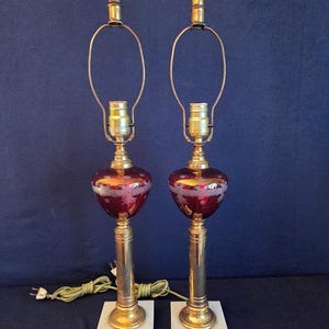 May include: Pair of vintage table lamps with ruby red glass globes, etched with a floral design. The lamps have brass accents, a white marble base, and a dark brown finial. The lamps are connected to a green electrical cord.