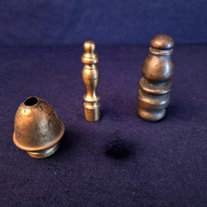 May include: Three antique brass objects are displayed against a dark blue background. The objects include a bell-shaped piece, a small, decorative spindle, and a cylindrical stopper with a rounded top. The brass has a warm, aged patina.