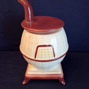 May include: A vintage ceramic stove-shaped cookie jar. The jar is primarily off-white with a maroon lid, handle, and base. It features a rectangular design with a yellow and white grid pattern.