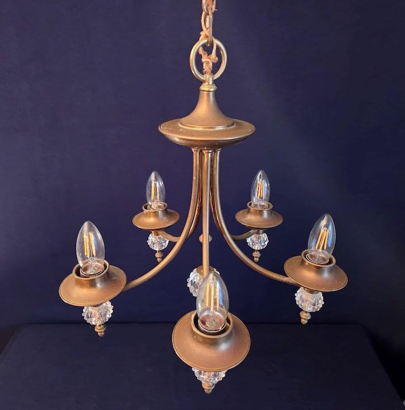 May include: A vintage gold-toned chandelier with five light fixtures. Each fixture has a bulb, a decorative crystal element, and a circular shade. The chandelier is suspended from a chain and has a central decorative element.