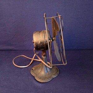 Peut inclure: Un ancien ventilateur &eacute;lectrique avec un bo&icirc;tier de moteur, des pales et une cage noirs. Le ventilateur a un socle d&eacute;coratif et un cordon d'alimentation. Il date probablement du d&eacute;but du XXe si&egrave;cle et montre des signes d'usure.