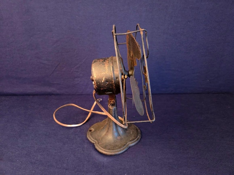 Peut inclure: Un ancien ventilateur &eacute;lectrique avec un bo&icirc;tier de moteur, des pales et une cage noirs. Le ventilateur a un socle d&eacute;coratif et un cordon d'alimentation. Il date probablement du d&eacute;but du XXe si&egrave;cle et montre des signes d'usure.