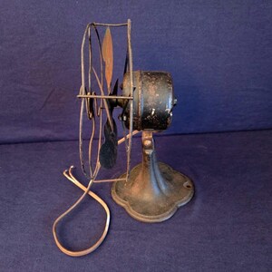 Peut inclure: Un ancien ventilateur &eacute;lectrique avec un bo&icirc;tier de moteur noir et une base d&eacute;corative. Les pales du ventilateur sont sombres et la cage de protection est en m&eacute;tal. Le cordon d'alimentation est visible et le ventilateur montre des signes d'usure.