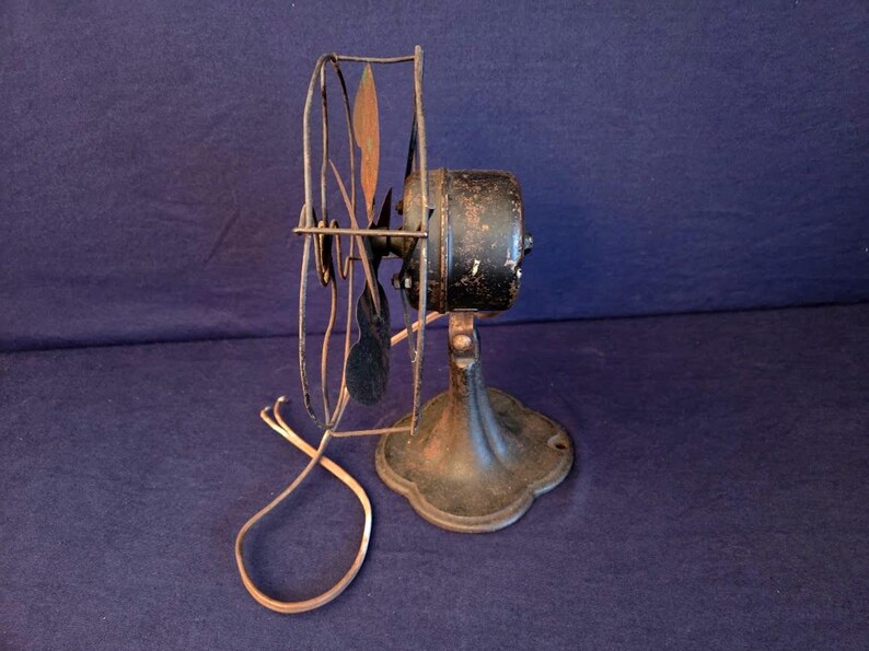 Peut inclure: Un ancien ventilateur &eacute;lectrique avec un bo&icirc;tier de moteur noir et une base d&eacute;corative. Les pales du ventilateur sont sombres et la cage de protection est en m&eacute;tal. Le cordon d'alimentation est visible et le ventilateur montre des signes d'usure.