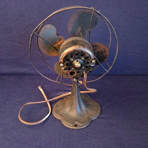 Peut inclure: Un ancien ventilateur &eacute;lectrique d'aspect sombre et vieilli. Le ventilateur a une cage circulaire, quatre pales et un bo&icirc;tier de moteur rond. Le socle est orn&eacute; et le cordon d'alimentation est visible. Le ventilateur est probablement en m&eacute;tal et pr&eacute;sente des traces de rouille.