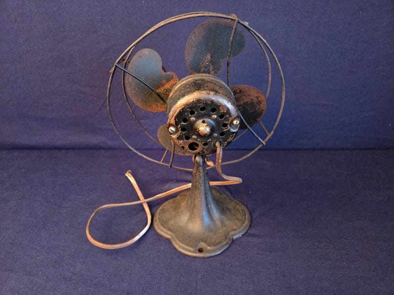 Peut inclure: Un ancien ventilateur &eacute;lectrique d'aspect sombre et vieilli. Le ventilateur a une cage circulaire, quatre pales et un bo&icirc;tier de moteur rond. Le socle est orn&eacute; et le cordon d'alimentation est visible. Le ventilateur est probablement en m&eacute;tal et pr&eacute;sente des traces de rouille.