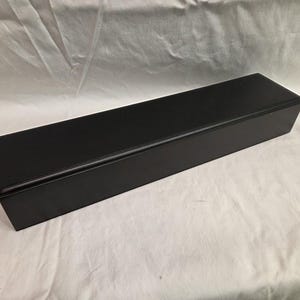 May include: A long, rectangular black box with a smooth, matte finish and a hinged lid. The box features clean, sharp edges and a solid black exterior, presenting a simple, elongated form designed for storage or display purposes.