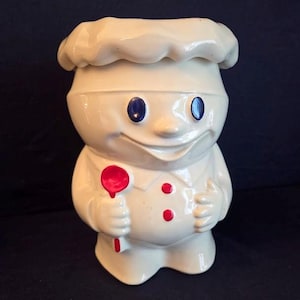 May include: A ceramic cookie jar in the shape of a smiling chef, wearing a white hat and uniform. The chef has blue eyes and holds a red spoon. Three red dots adorn the chef's uniform.