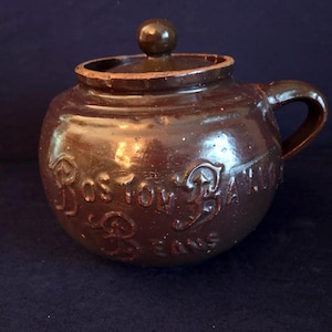 May include: A brown ceramic bean pot with a lid and handle. The words "Boston Baked Beans" are embossed on the side. The pot has a round shape and a dark brown glaze.