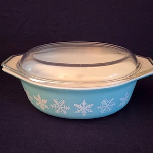 May include: A vintage Pyrex casserole dish with a clear glass lid. The dish is light blue with white snowflake designs around the rim. The lid is clear, and the handles are white with gold trim. A classic kitchenware item.