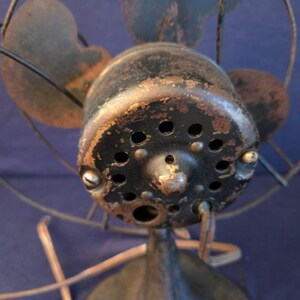 Peut inclure: Gros plan d'un ventilateur &eacute;lectrique ancien. Le bo&icirc;tier du moteur noir pr&eacute;sente des signes d'usure et de vieillissement, avec plusieurs petits trous. Les pales du ventilateur sont d'une couleur sombre et sourde, et la cage m&eacute;tallique est rouill&eacute;e.