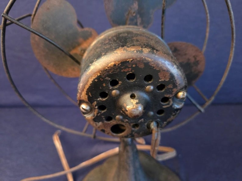Peut inclure: Gros plan d'un ventilateur &eacute;lectrique ancien. Le bo&icirc;tier du moteur noir pr&eacute;sente des signes d'usure et de vieillissement, avec plusieurs petits trous. Les pales du ventilateur sont d'une couleur sombre et sourde, et la cage m&eacute;tallique est rouill&eacute;e.